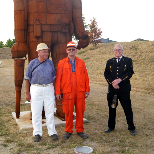 Vor der Stature eines Bergmann stehen Drei Bergleute in Ihrer Arbeitskleidung.